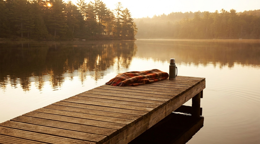 a wooden dock reaching into a misty lake at golden hour, with a folded plaid blanket and a thermos at the end